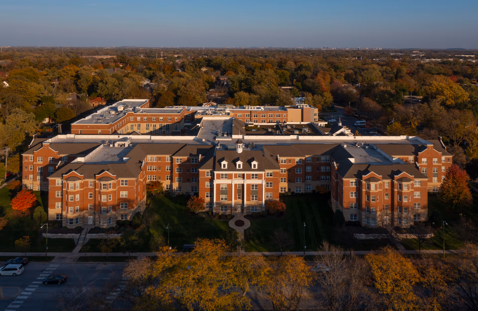 Aerial view of a large multi-story brick building with white accents, surrounded by trees with autumn foliage. The building appears to be a senior living facility with a well-maintained lawn and pathways in front. The sky is clear and the surrounding area is filled with dense trees and residential homes.