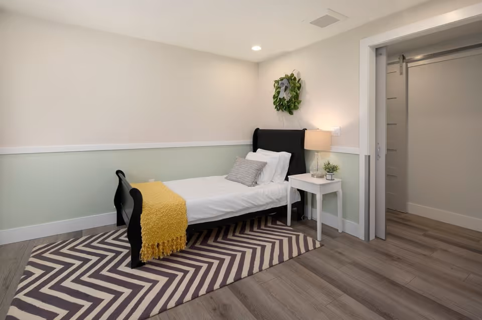 A small bedroom with a single black bed frame, white bedding, a gray pillow, and a yellow throw blanket. There is a white nightstand with a lamp and a small plant next to the bed. A green wreath hangs on the wall above the bed. The room has light-colored walls with a white and light green trim and wood flooring with a chevron-patterned rug. A sliding barn door is partially open, revealing a hallway.