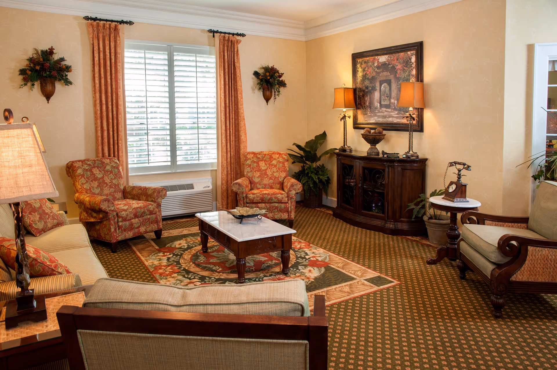 A cozy living room with patterned carpet and floral upholstery. The room features two floral armchairs, a beige sofa, a wooden coffee table with a marble top, and a wooden sideboard with two lamps and a decorative bowl. There are orange curtains framing a window with white blinds, wall-mounted floral arrangements, a framed painting, and a vintage-style telephone on a small round side table.