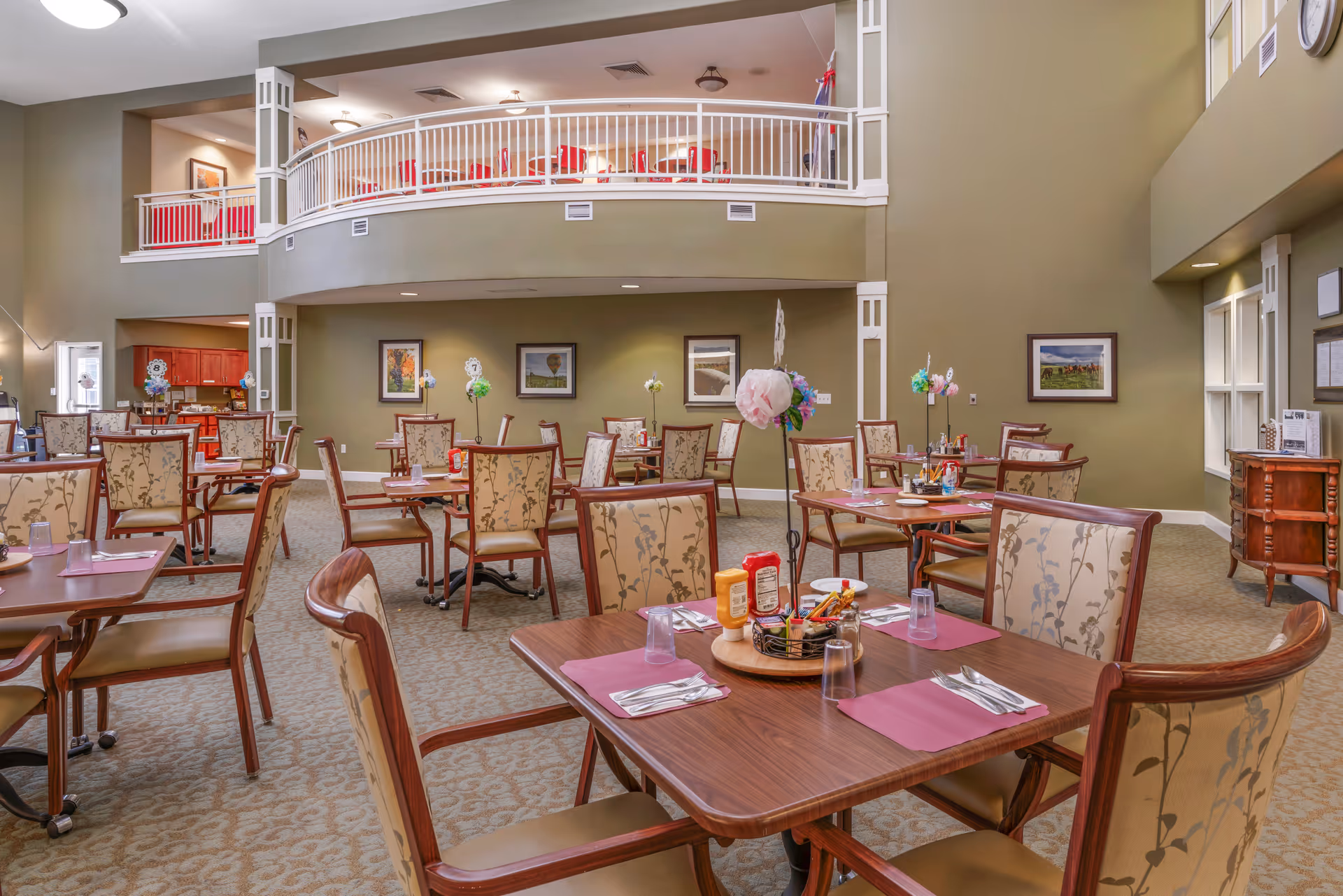 Dining room with multiple set tables and upholstered chairs beneath a second-floor balcony in a senior living facility.