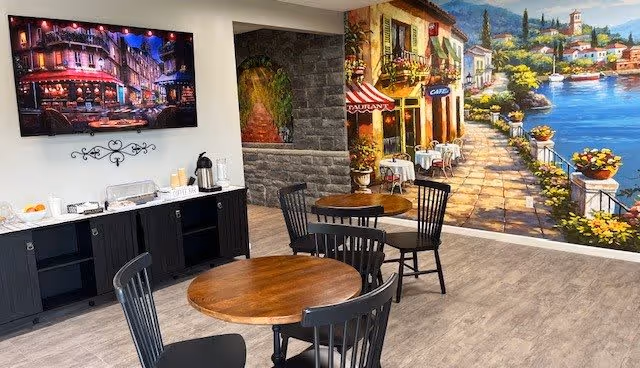 Small communal dining area with round wooden tables and black chairs, a buffet counter and a large mural of a seaside village on the wall.