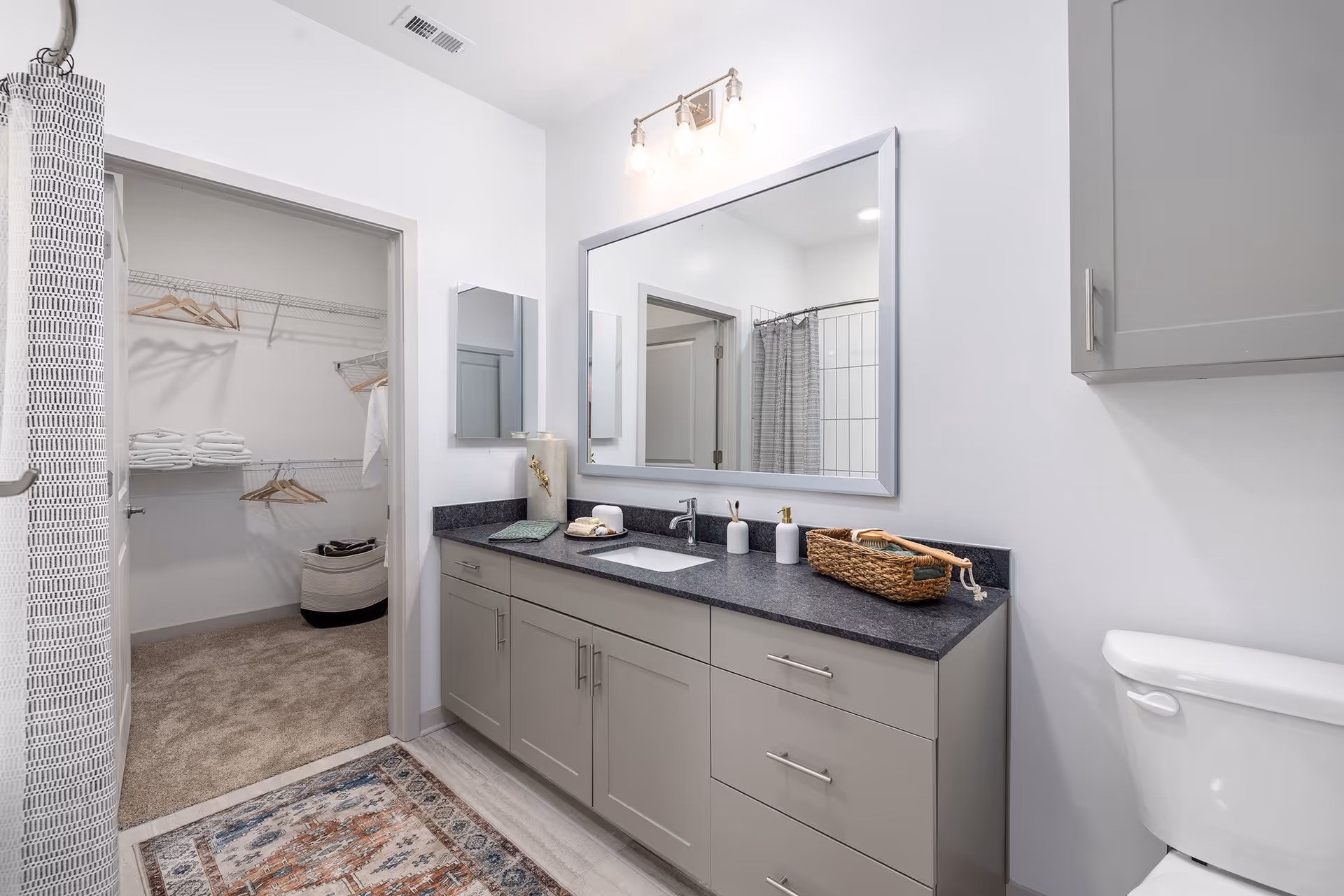 A modern bathroom with a large mirror above a gray vanity with a black countertop. The vanity has a sink, soap dispensers, a basket, and decorative items. To the right is a white toilet, and to the left is a doorway leading to a walk-in closet with shelves, hangers, and folded towels. A shower curtain is partially visible on the left side of the image.