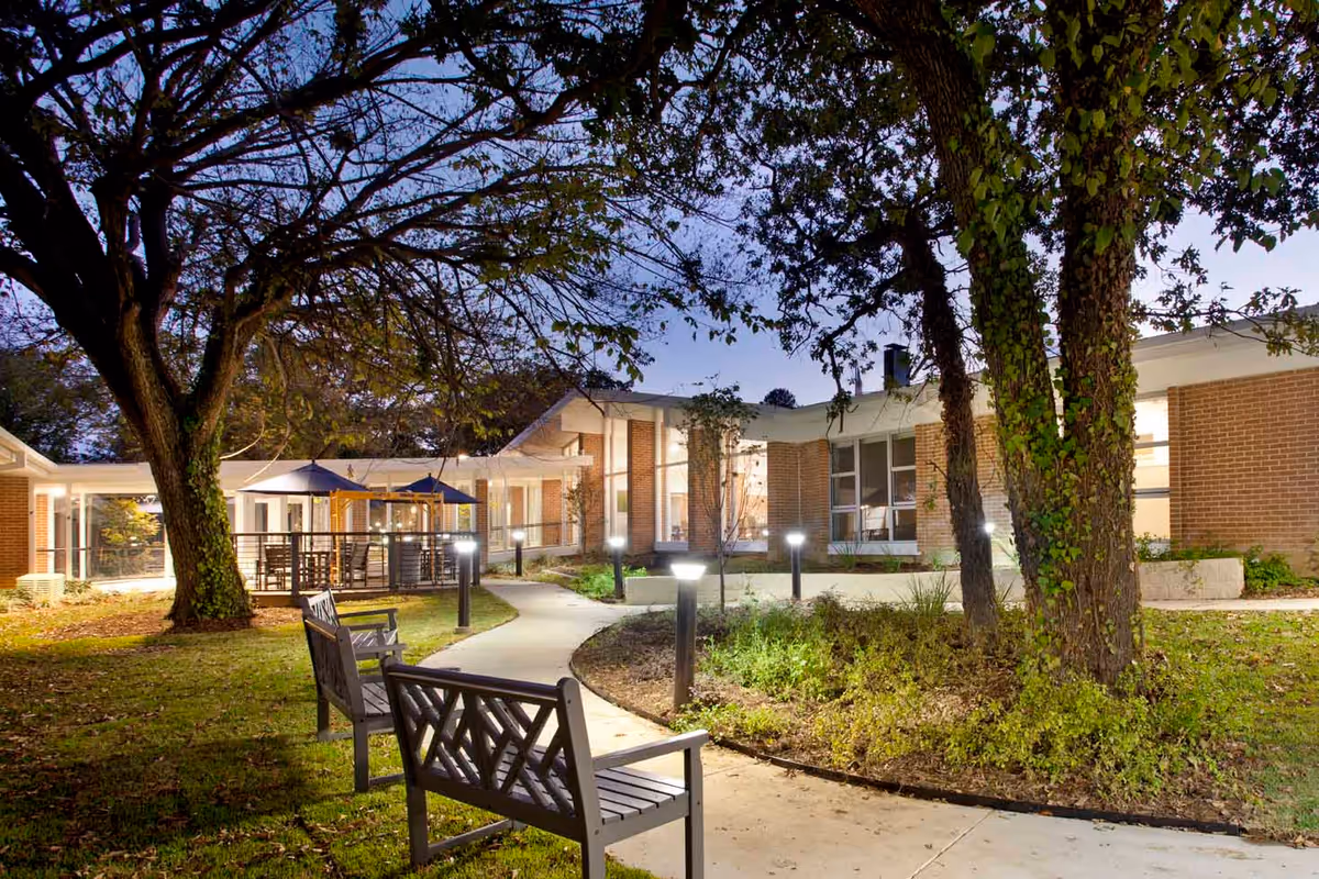 Outdoor view of a senior living facility courtyard at dusk with a curved concrete pathway, benches, trees, and illuminated lamp posts. The building has large windows and brick walls, and there are patio umbrellas visible in the background.