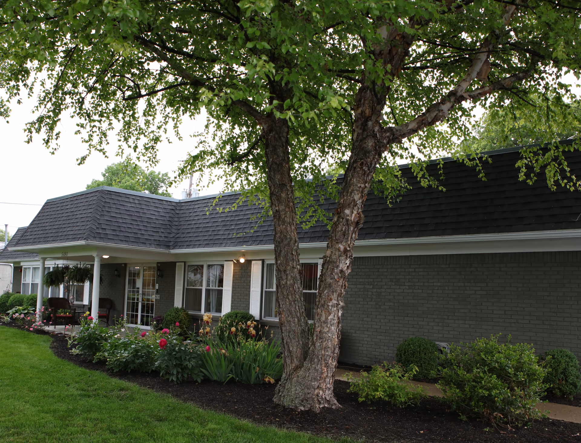 Exterior view of a single-story brick building with a dark shingled roof, surrounded by a well-maintained garden with green shrubs, flowering plants, and a large tree in the foreground. There is a covered porch area with chairs near the entrance.