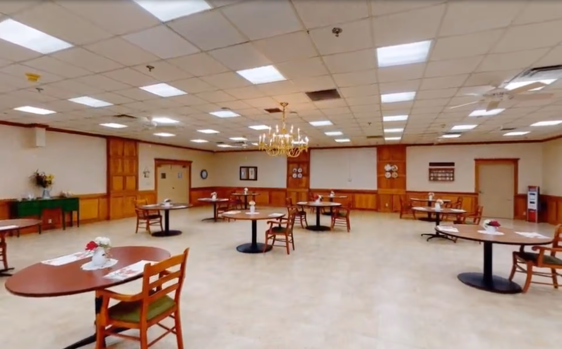 A spacious dining room with multiple round tables and wooden chairs arranged evenly throughout the room. Each table has a small floral centerpiece and placemats. The room features a tiled floor, wood-paneled lower walls, cream-colored upper walls, and a ceiling with fluorescent lighting and a chandelier in the center. There are doors and decorative plates on the walls.