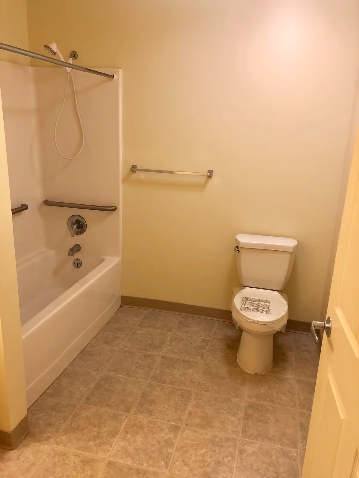 A simple beige bathroom showing a bathtub/shower with grab bars, a towel bar, and a toilet with a plastic cover.