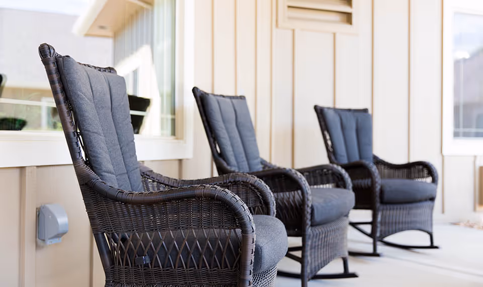 Three dark wicker rocking chairs with gray cushions lined up on a covered porch outside a light-colored building.