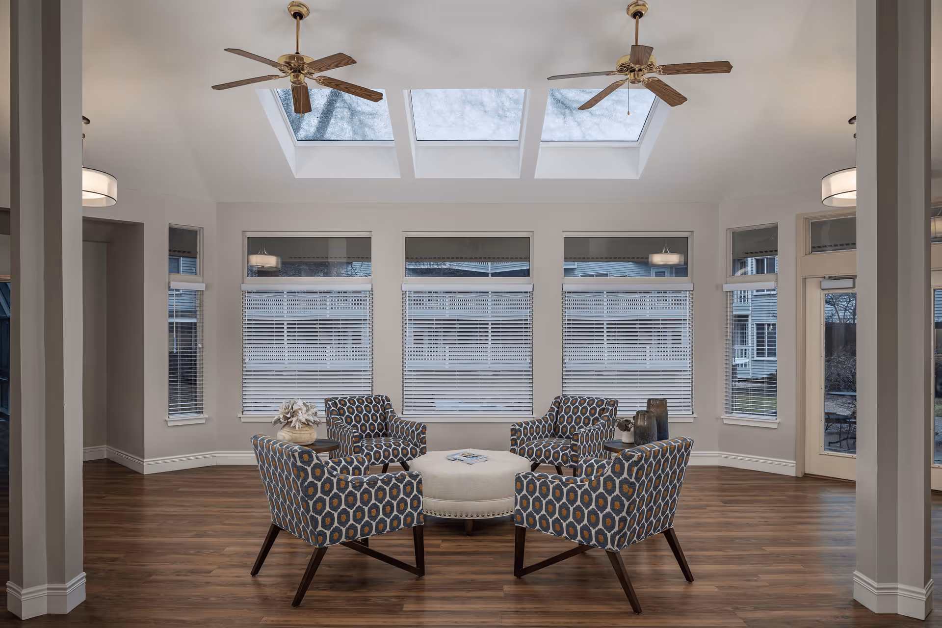 A bright and spacious sitting area with four patterned armchairs arranged around a round ottoman. The room features large windows with blinds, three skylights in the ceiling, two ceiling fans, and wood flooring. The walls are painted light gray, and there are two pendant lights visible in adjacent rooms.