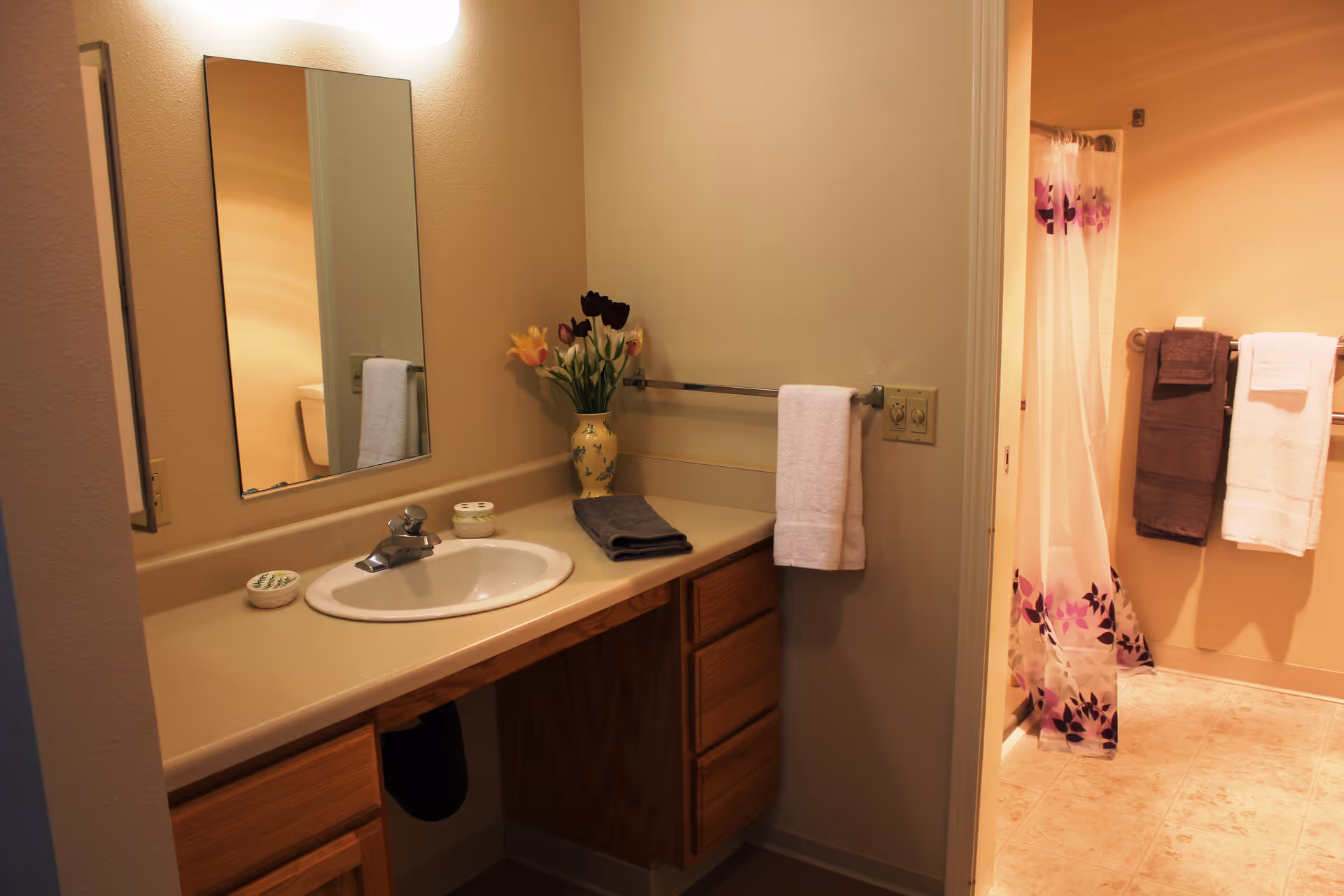 A bathroom vanity with a sink, faucet, and a large mirror above it. On the countertop, there is a vase with flowers, a folded dark towel, and two small containers. A white towel hangs on a towel rack attached to the wall. In the background, there is a shower area with a floral-patterned shower curtain and two towels hanging on a rack.