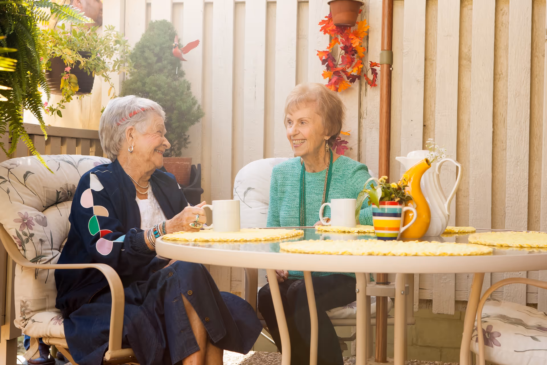 Two elderly women sitting at a round outdoor table with cushioned chairs, enjoying a conversation and smiling. The table has a white pitcher, a yellow pitcher, a colorful striped mug, and a small plant. The setting is a cozy patio with plants and autumn decorations on the wall behind them.