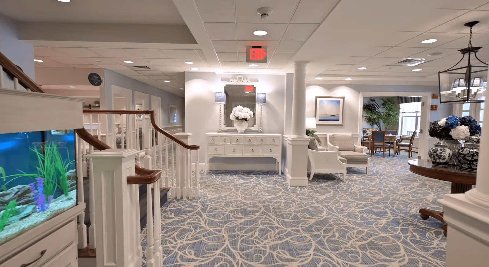 Interior view of a senior living facility lobby area with a patterned blue and beige carpet, white furniture including chairs and a console table with a mirror and lamps, a large fish tank on the left, a round table with blue and white floral arrangements on the right, and a dining area visible in the background.