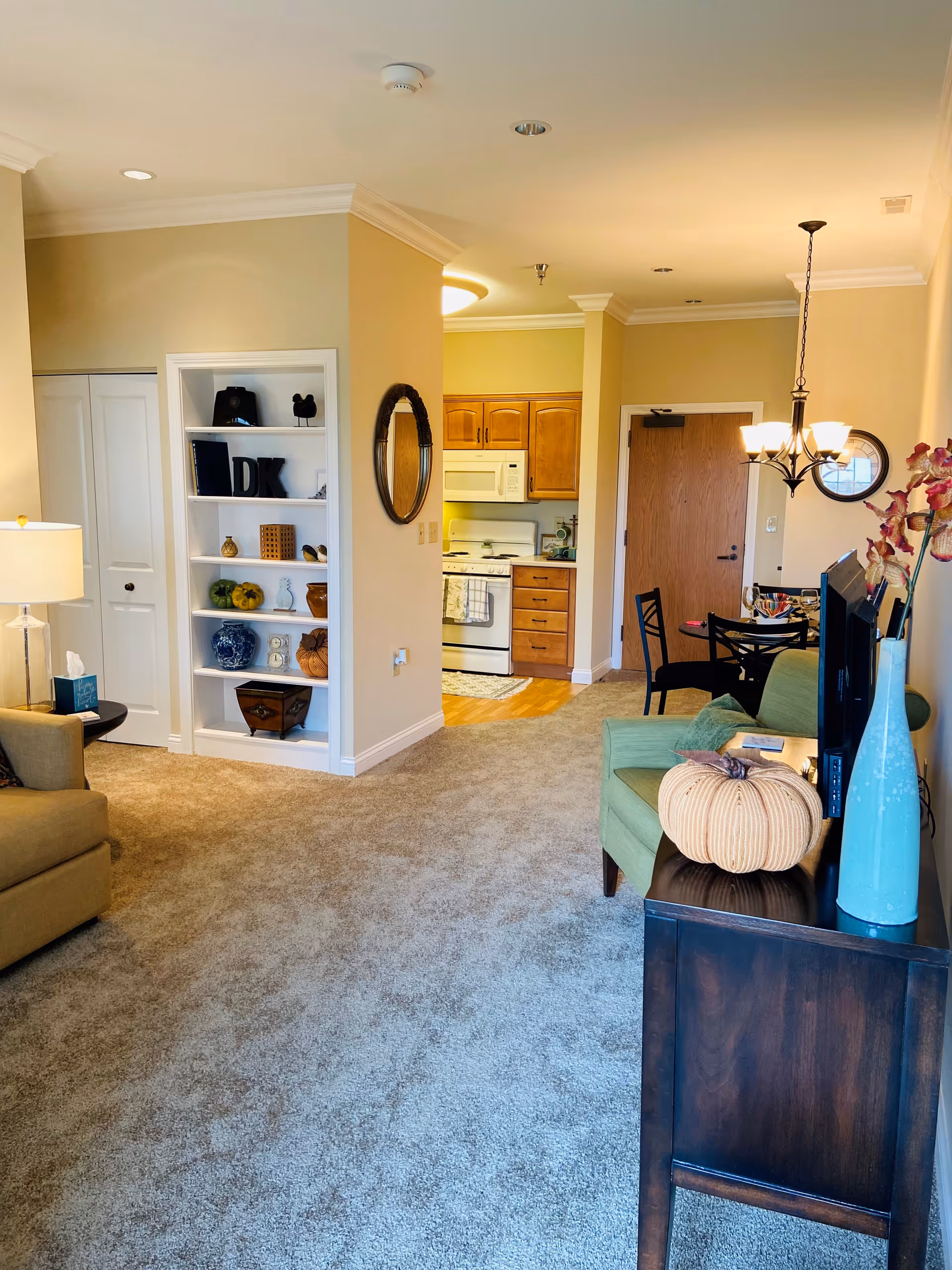 Carpeted open living room with armchairs, a TV console and decorative shelving, looking toward a small kitchen and dining area.