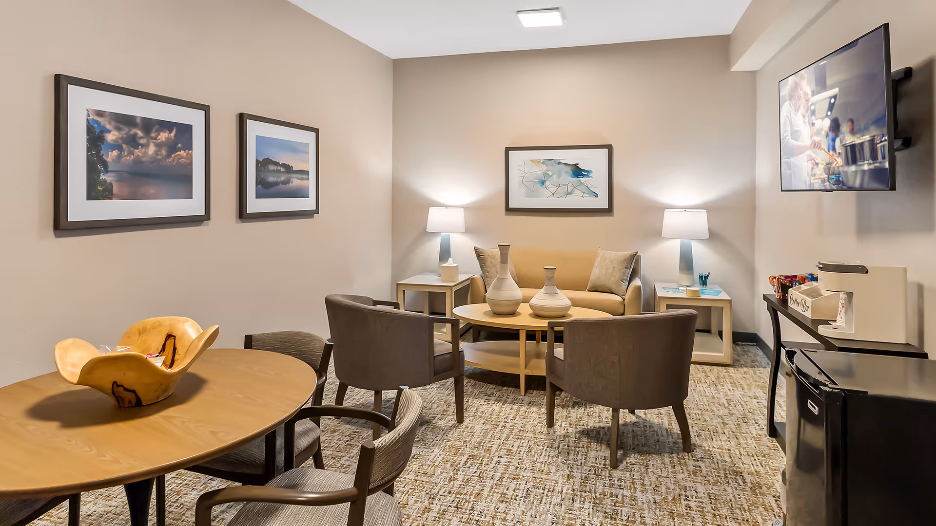 A cozy living room area with a beige couch, two brown armchairs, a round wooden coffee table with decorative vases, two side tables with lamps, a wall-mounted TV showing a cooking scene, a wooden dining table with four chairs, and three framed pictures on the walls.