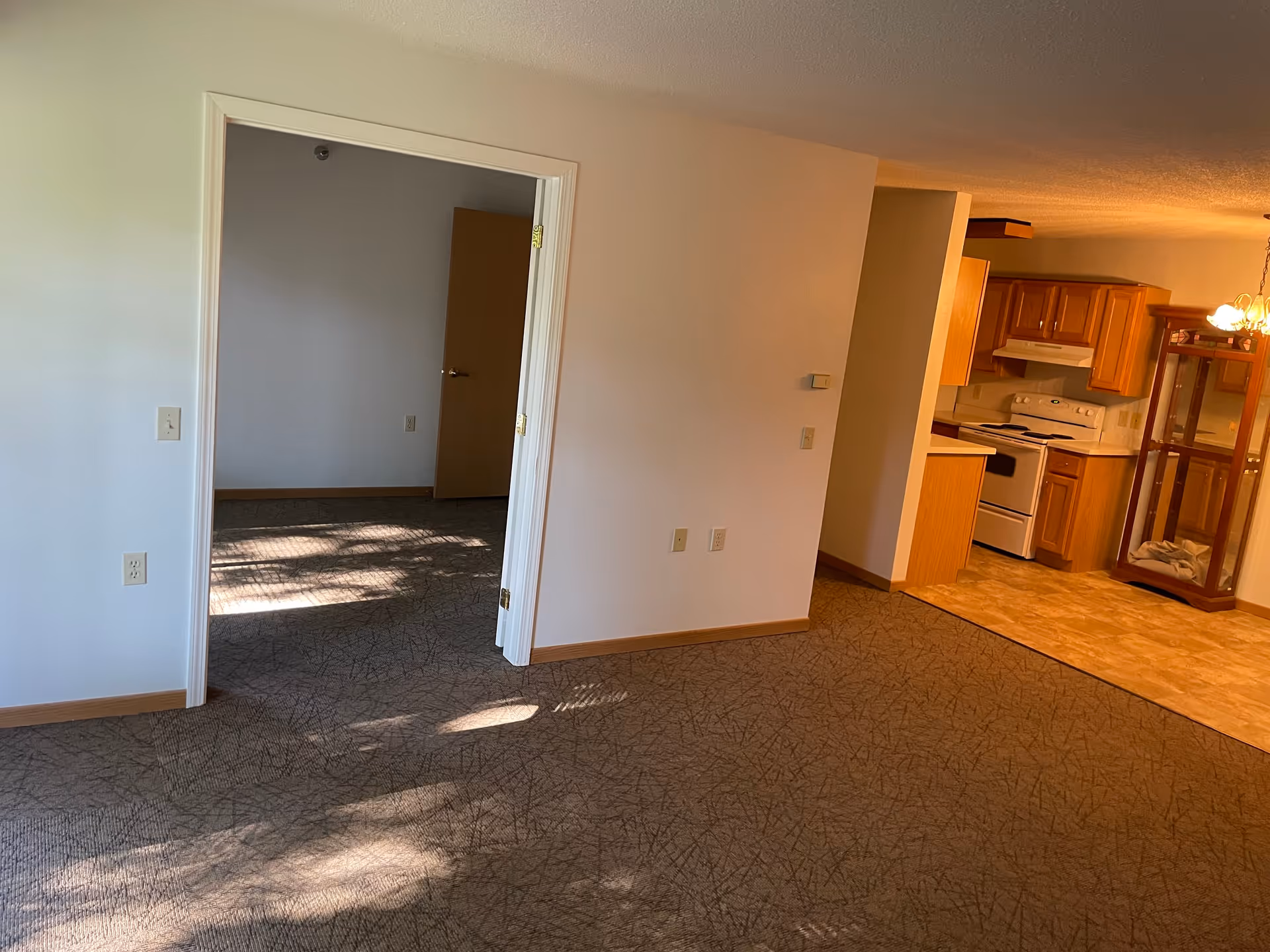 Interior view of a senior living facility showing a carpeted living area leading to a kitchen with wooden cabinets and white appliances. There is an open doorway to another room with carpeted flooring and a closed wooden door. The walls are painted light beige and there is a glass display cabinet near the kitchen area.