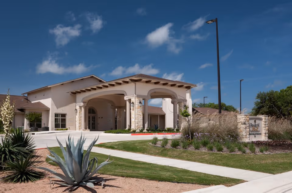 Exterior view of Franklin Park Boerne senior living facility showing the entrance with a covered driveway, landscaped garden with desert plants, and a clear blue sky.