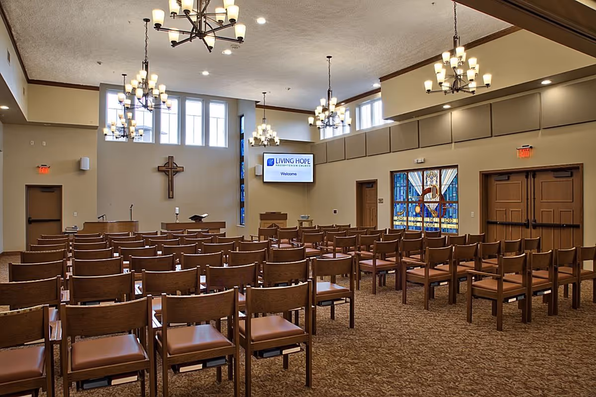 Interior view of a chapel or worship room with rows of wooden chairs facing a podium and a cross mounted on the wall. The room has multiple chandeliers hanging from the ceiling, large windows letting in natural light, and a screen displaying 'Living Hope Presbyterian Church Welcome'. There is also a stained glass window and double wooden doors on the right side.