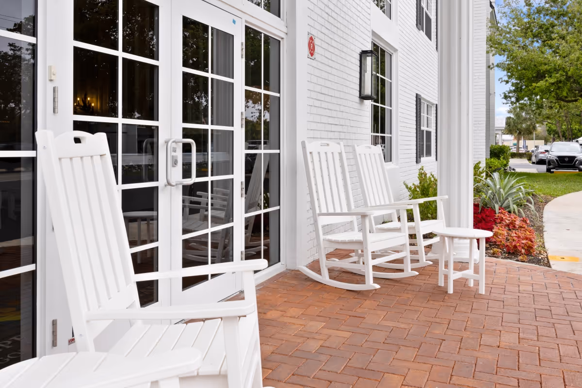 White rocking chairs and a small white table on a brick patio outside a white building with large glass doors and windows, surrounded by greenery and parked cars in the background.