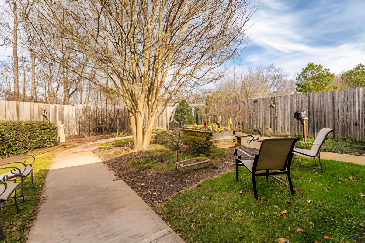 Outdoor garden area with a paved walkway, green grass, leafless trees, bushes, and several benches for seating. A wooden fence encloses the space under a partly cloudy sky.