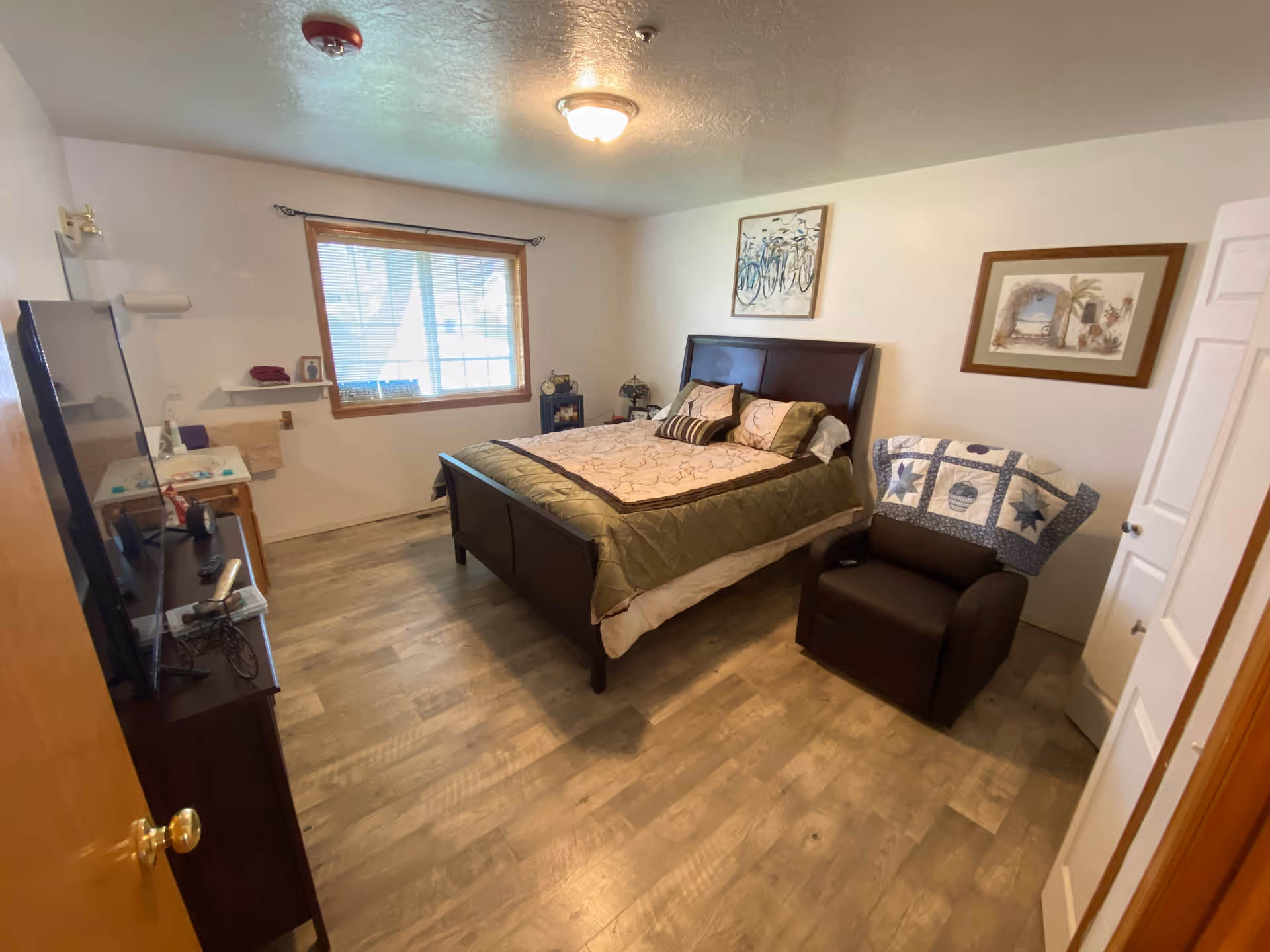 A cozy bedroom in Willow Place Assisted Living featuring a large bed with a green and beige quilt, a dark brown armchair with a quilt draped over the back, a flat-screen TV on a stand, a window with blinds, and framed artwork on the walls. The room has wooden flooring and a ceiling light fixture.