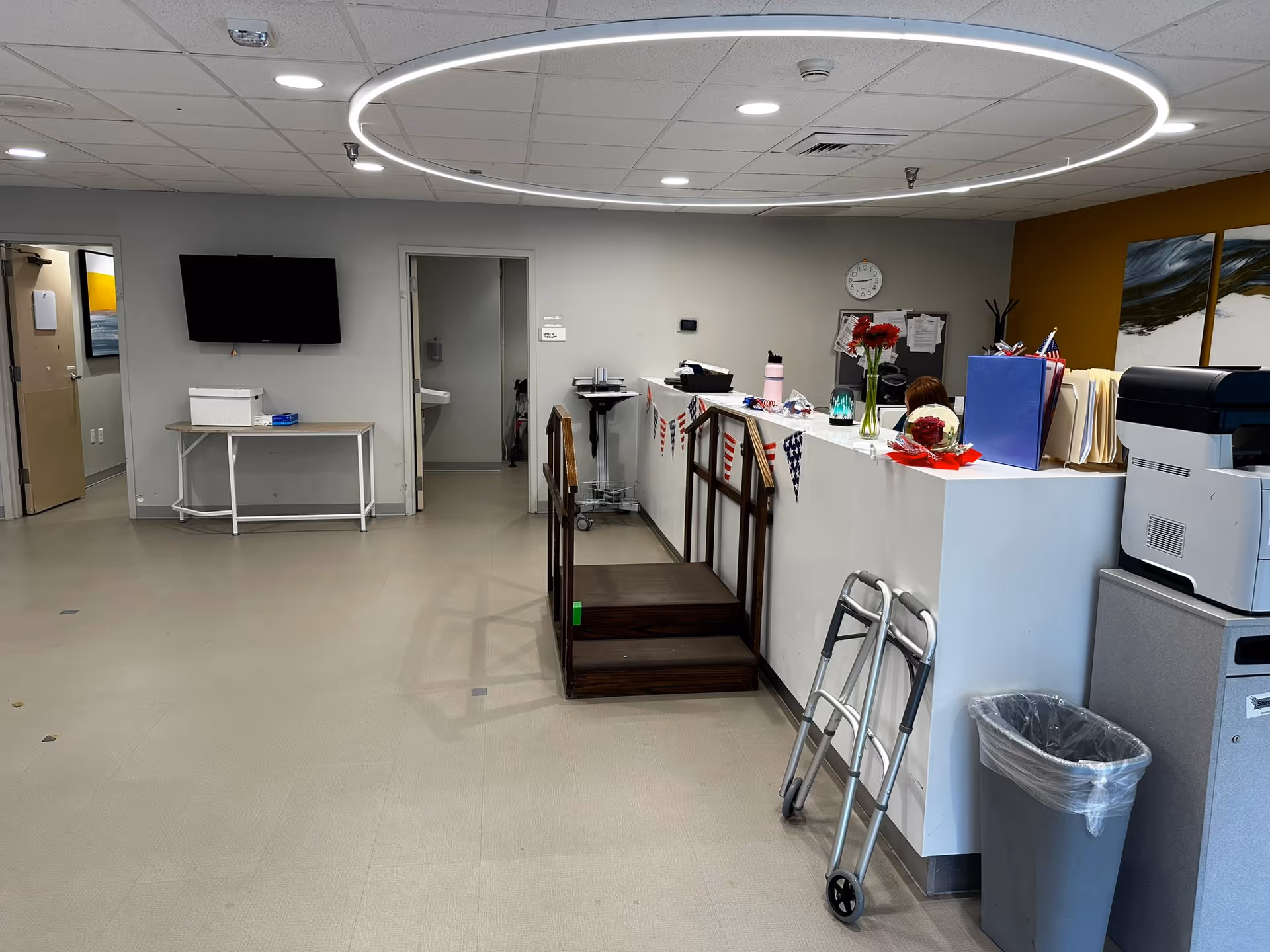Interior view of a nursing and rehabilitation center reception area with a white counter decorated with small American flag bunting. A walker is leaning against the counter next to a trash can and a printer. Behind the counter, a person is partially visible. The room has a beige floor, gray walls, a mounted TV, and a circular ceiling light fixture. There is a small wooden ramp leading up to the counter and an open door revealing a sink in a small room.