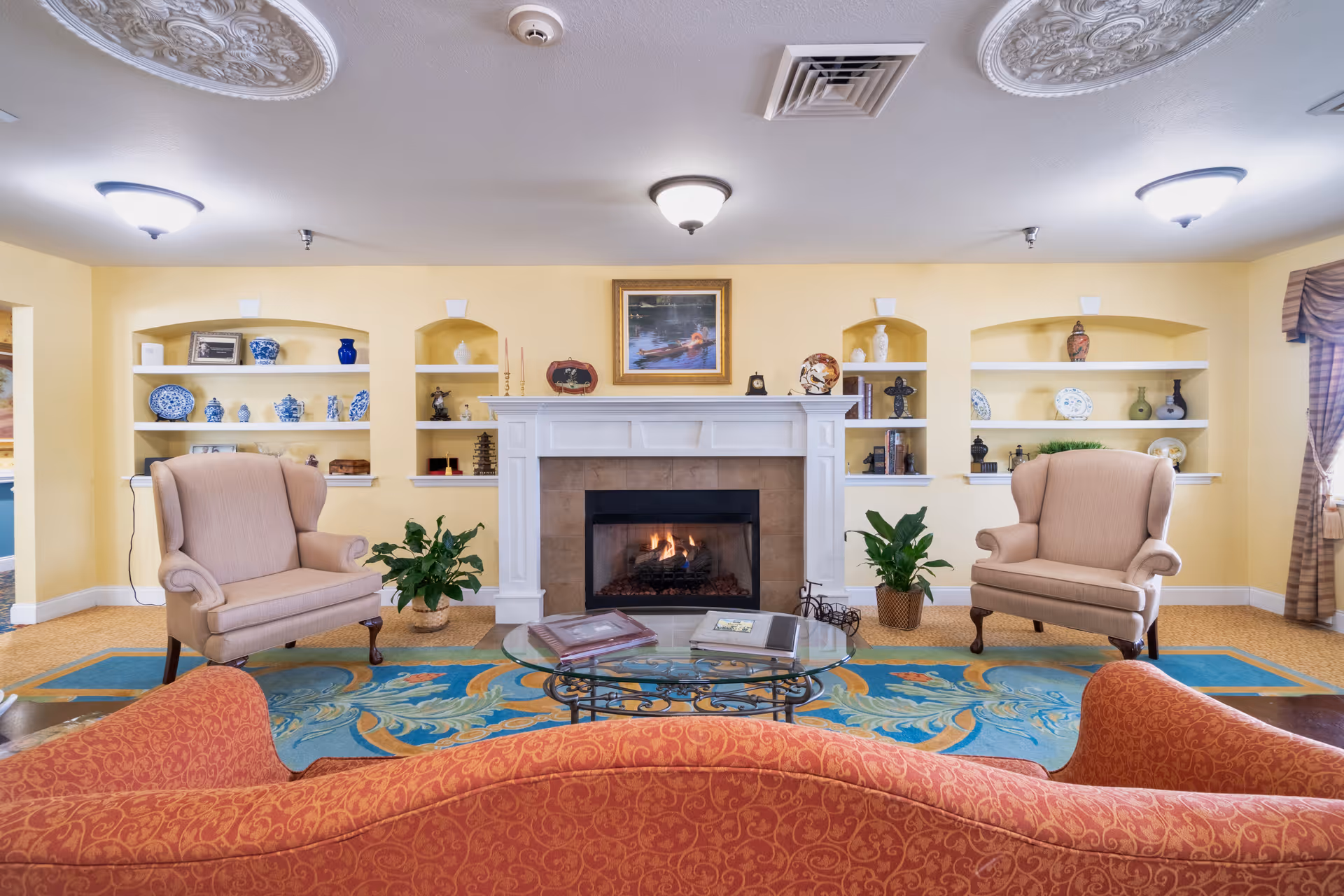 Cozy living room with a central fireplace, built-in shelves, two armchairs, a red sofa, glass coffee table and a colorful rug.