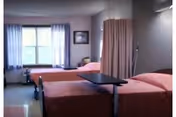 A nursing facility room with two hospital beds separated by a privacy curtain. Each bed has a pink bedspread and an overbed table. There is a window with curtains letting in natural light and a framed picture on the wall.