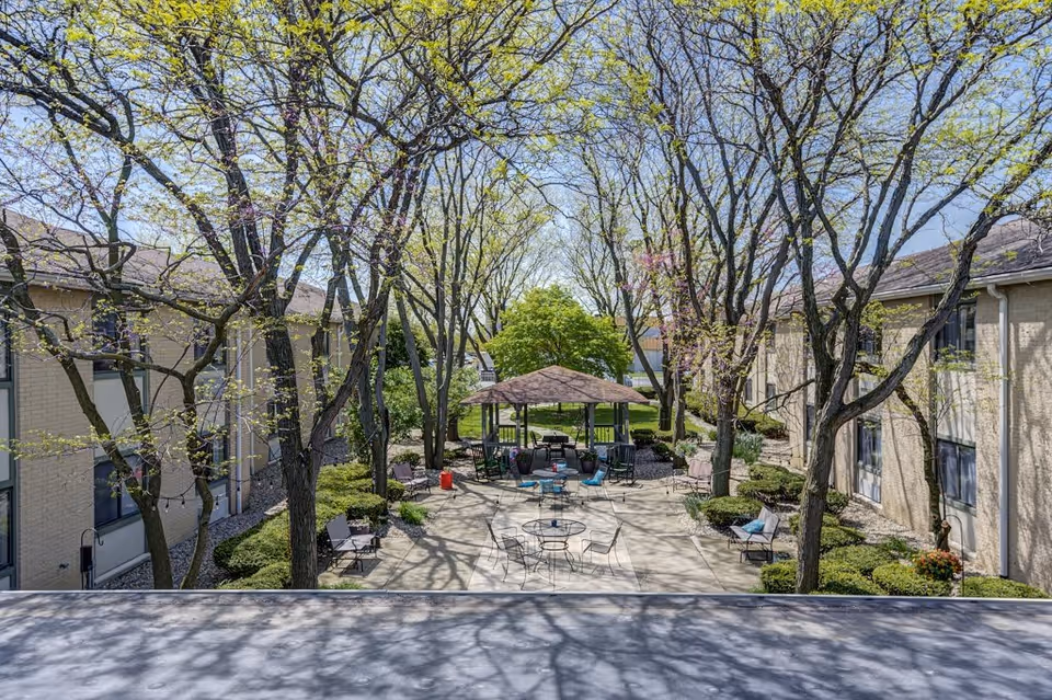 Outdoor courtyard area at The Commons A ProMedica Senior Living Community with leafing trees, patio tables and chairs, benches, and a gazebo in the center. The courtyard is surrounded by two beige buildings with windows.