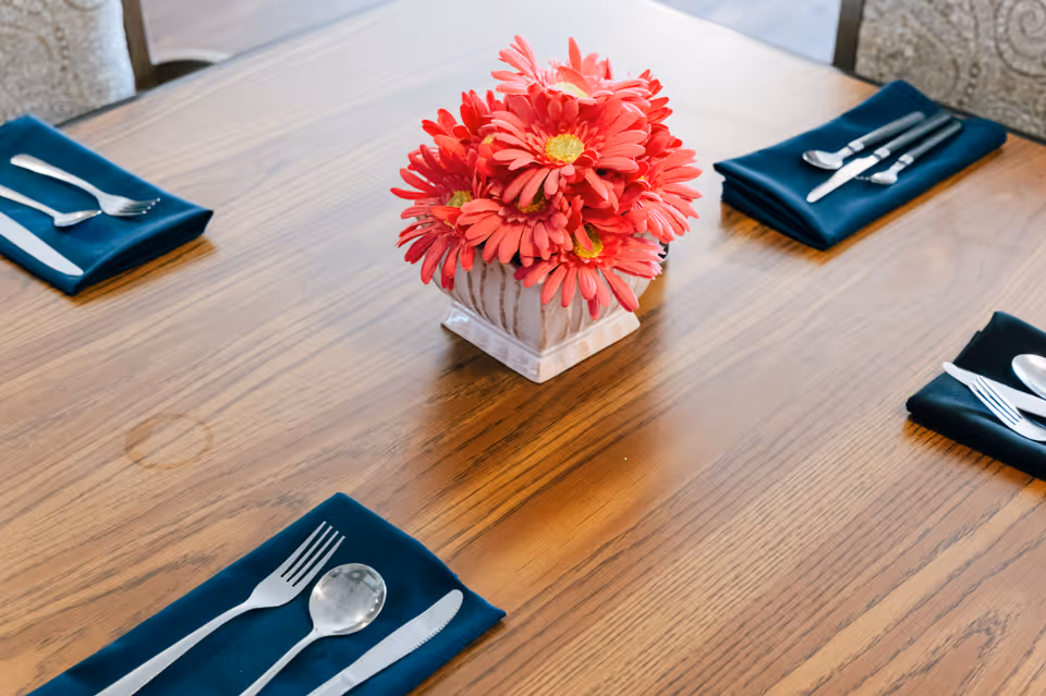 A wooden dining table set with four place settings, each with a fork, spoon, and knife on a dark blue napkin. A small white vase with bright pink flowers is placed in the center of the table.