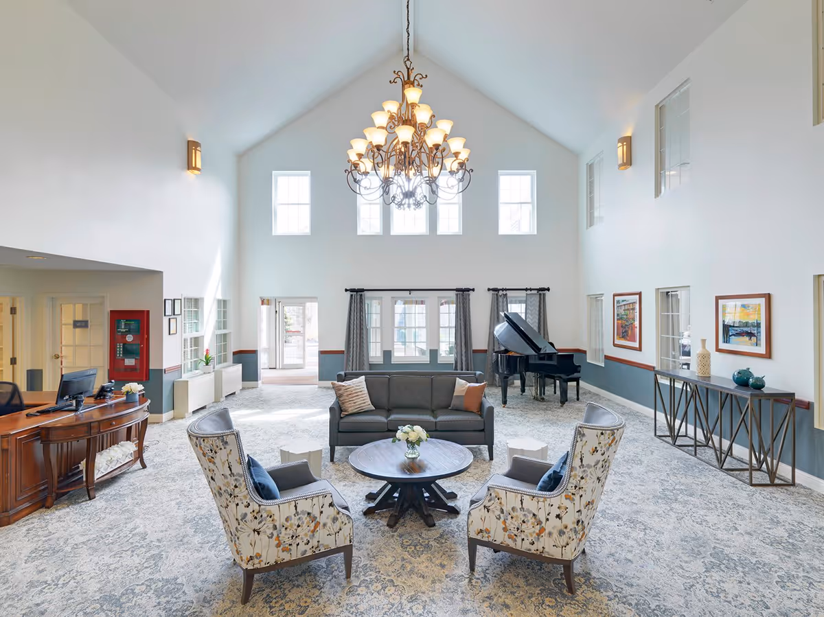 A spacious and well-lit common area with high vaulted ceilings and a large chandelier. The room features a gray sofa, two floral-patterned armchairs, and a round wooden coffee table with a flower vase. There is a grand piano in the corner, framed artwork on the walls, and a wooden reception desk to the left. Large windows and glass doors allow natural light to fill the space.