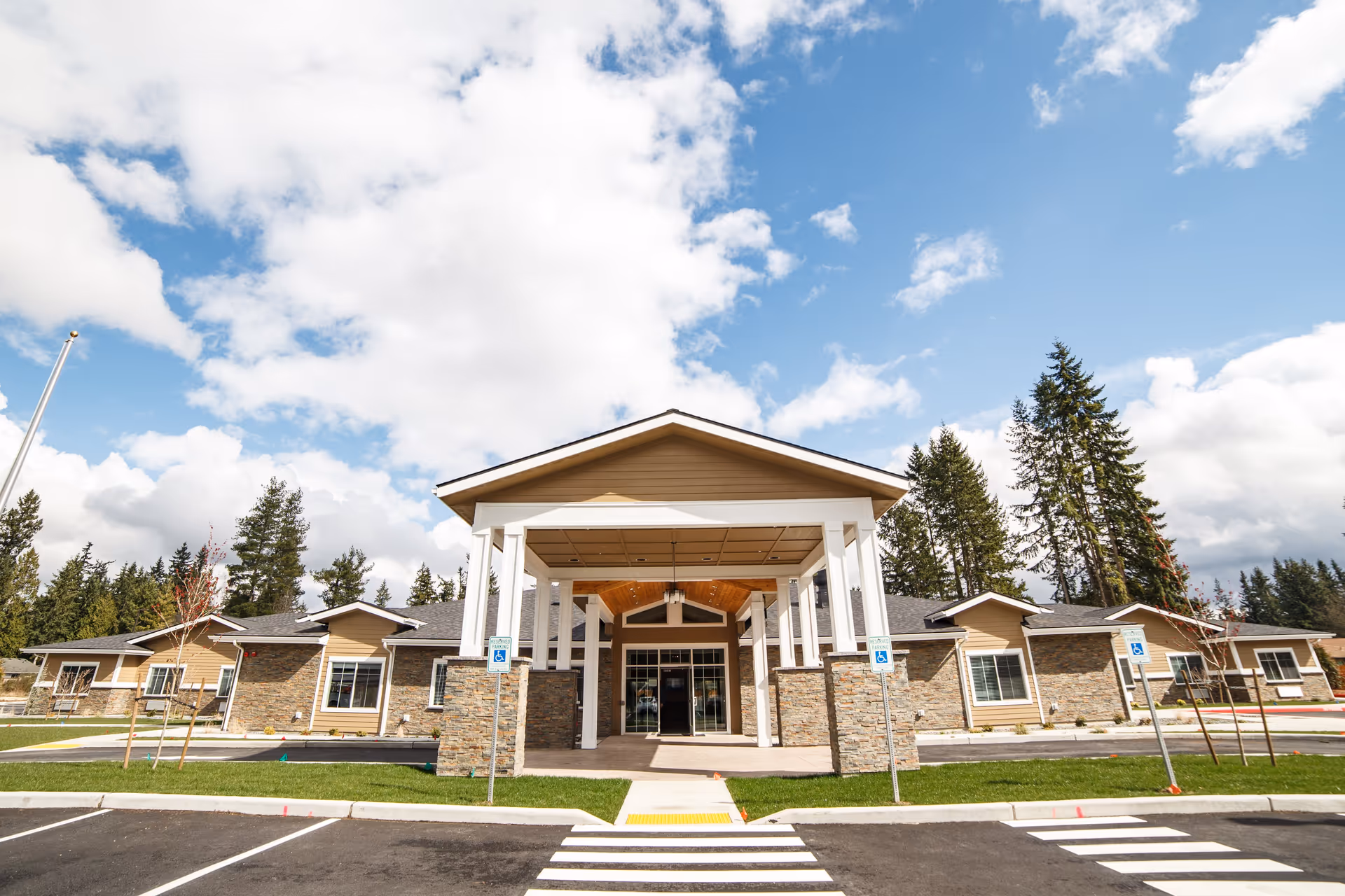 Front exterior view of Fieldstone Memory Care Marysville building with a covered entrance, stone and beige siding, surrounded by trees under a partly cloudy sky.
