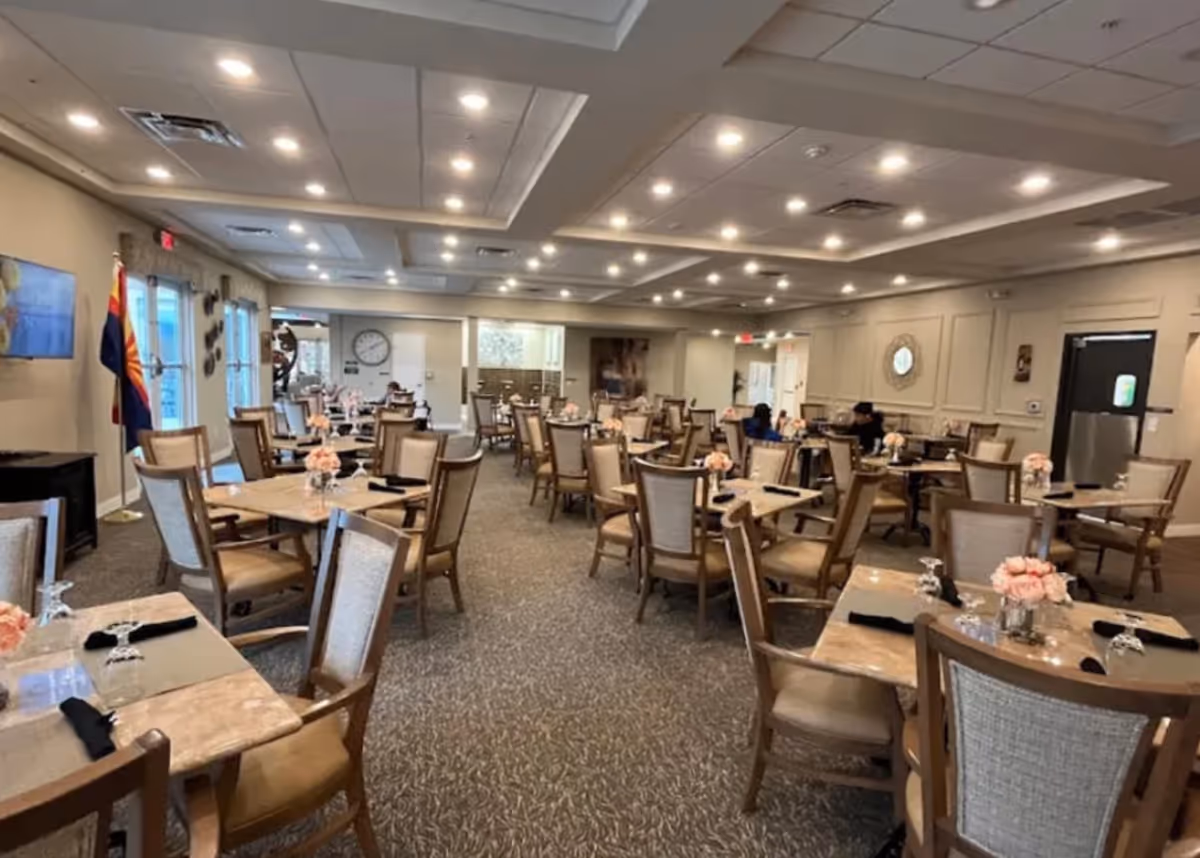 A spacious dining room with multiple tables and chairs arranged neatly. Each table has a small floral centerpiece and place settings with black napkins and upside-down glasses. The room is well-lit with recessed ceiling lights and has a carpeted floor. There are large windows on one side and a clock on the far wall. A few people are seated at tables in the background.