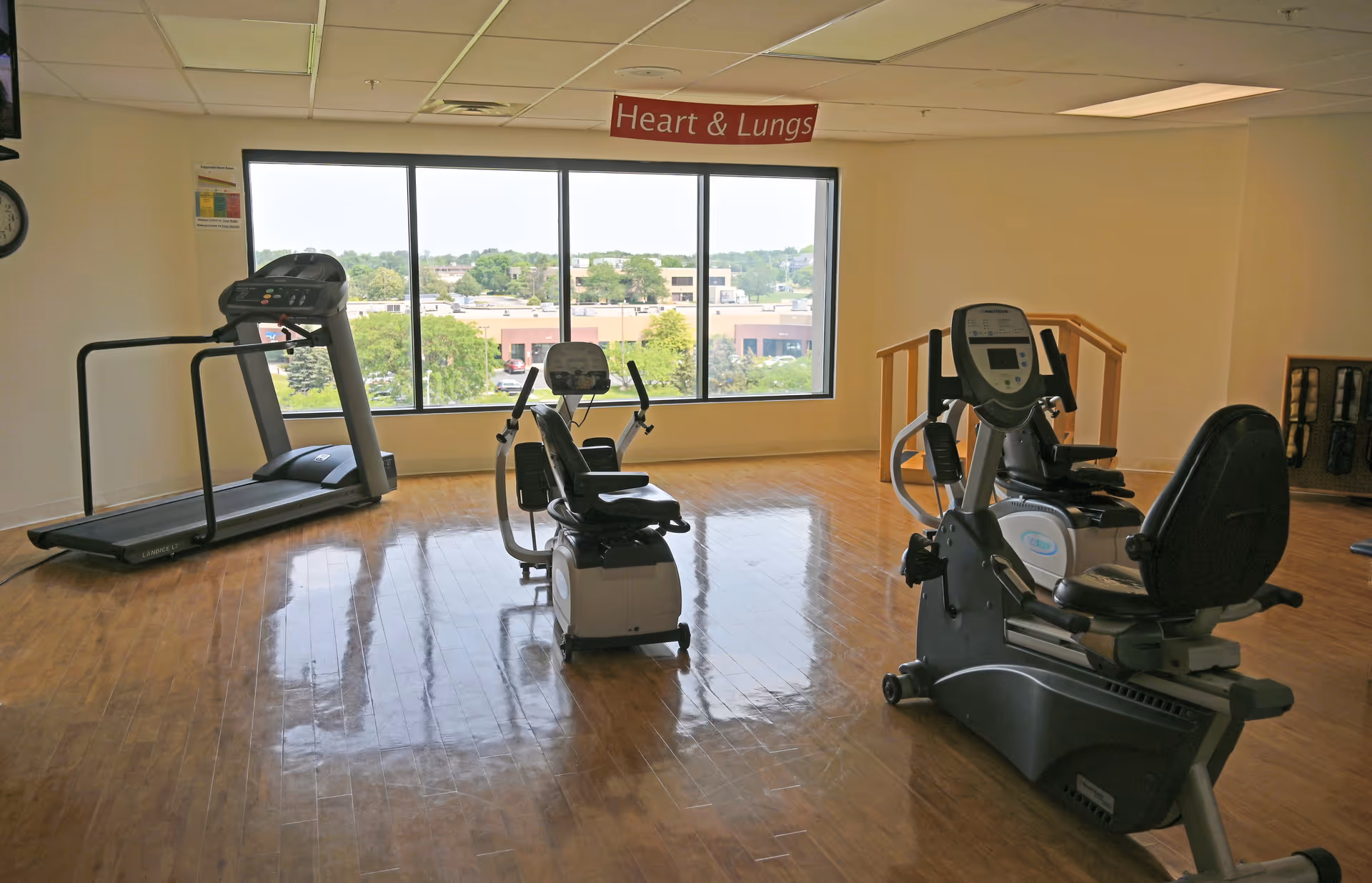 A fitness room with exercise equipment including a treadmill and two recumbent stationary bikes. The room has wooden flooring and a large window showing an outside view of buildings and trees. A sign above the window reads 'Heart & Lungs'.