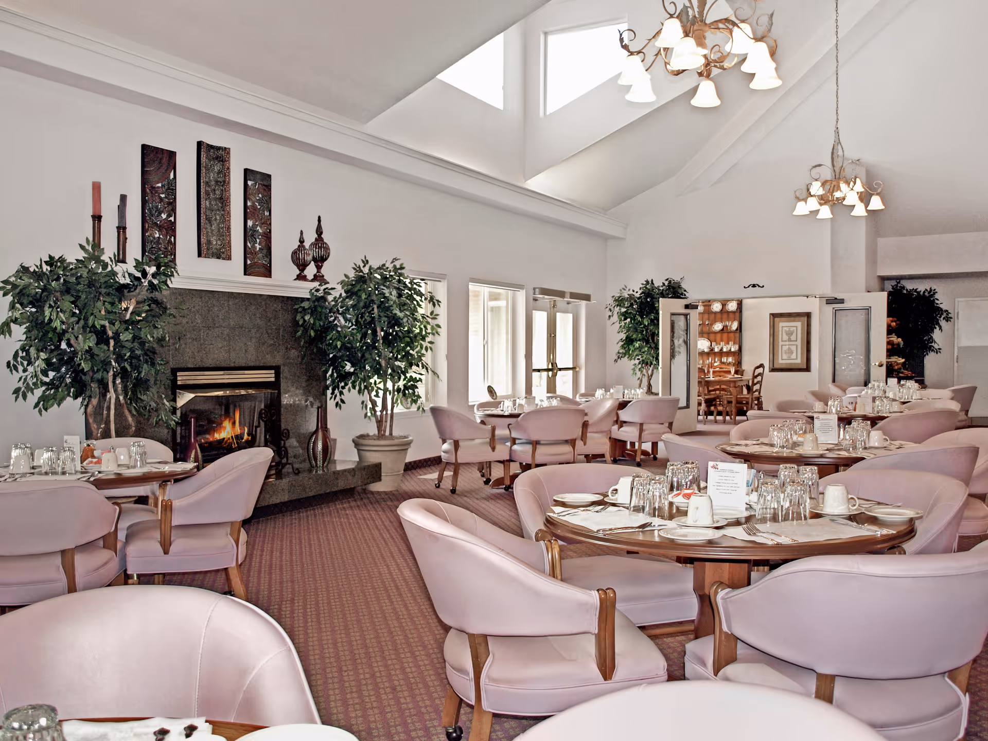 A spacious dining room with round wooden tables set with white cups, plates, and glasses. The room features lavender upholstered chairs, a fireplace with decorative items on the mantel, large potted plants, and high ceilings with multiple chandeliers. There are windows and double doors letting in natural light, and a cabinet with dishes is visible in the background.