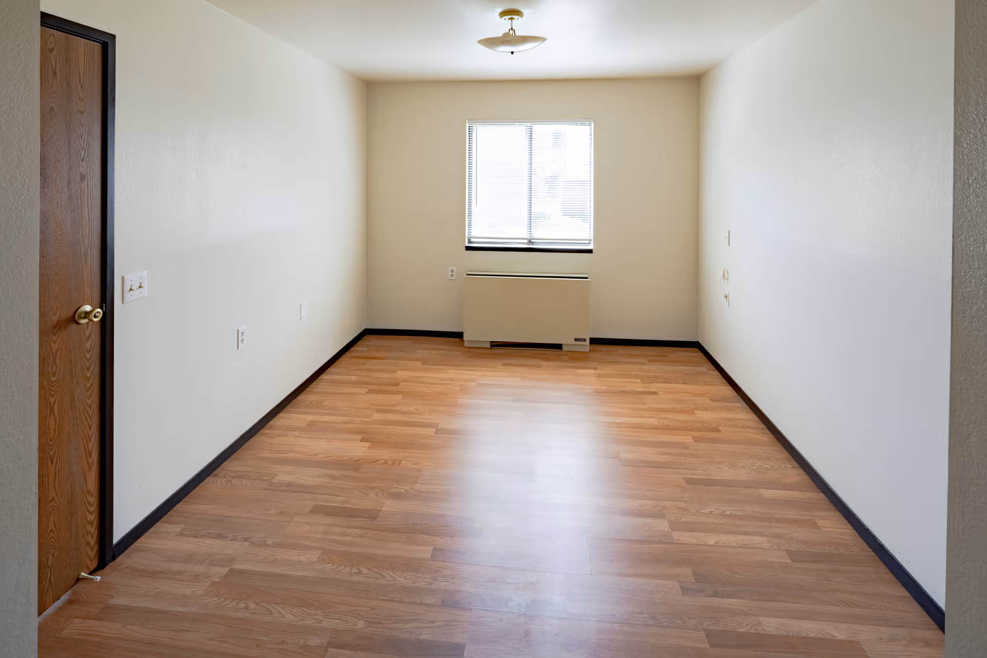 Empty rectangular room with wood laminate flooring, a window and wall heater at the far end and a closed wooden door on the left.