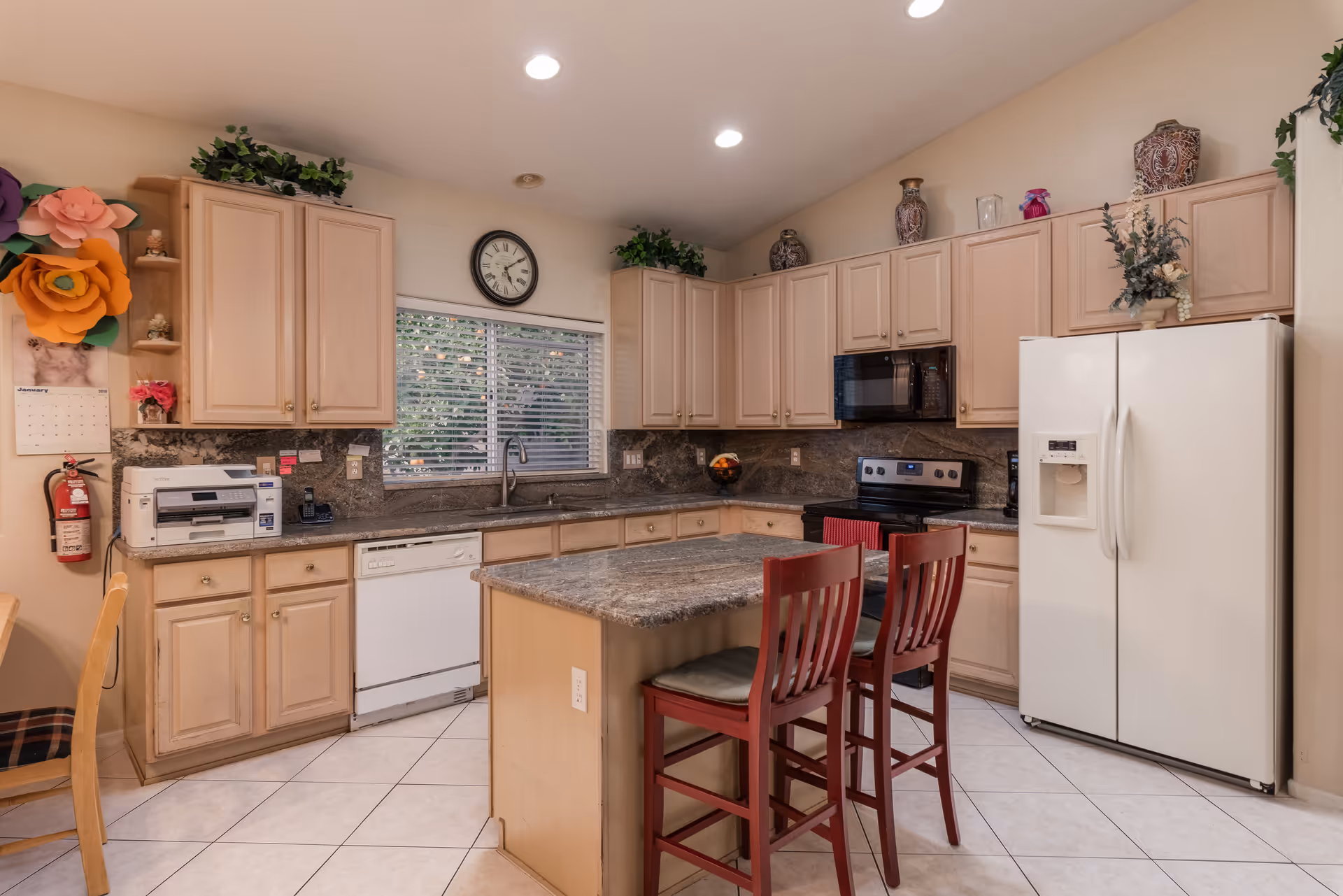 A kitchen with light wood cabinets, a granite countertop island with two red cushioned chairs, a white refrigerator with water dispenser, a black microwave above a black stove, a dishwasher, a sink under a window with blinds, and decorative plants and vases on top of the cabinets. There is a clock on the wall and a fire extinguisher mounted on the left side near a calendar and large paper flowers.
