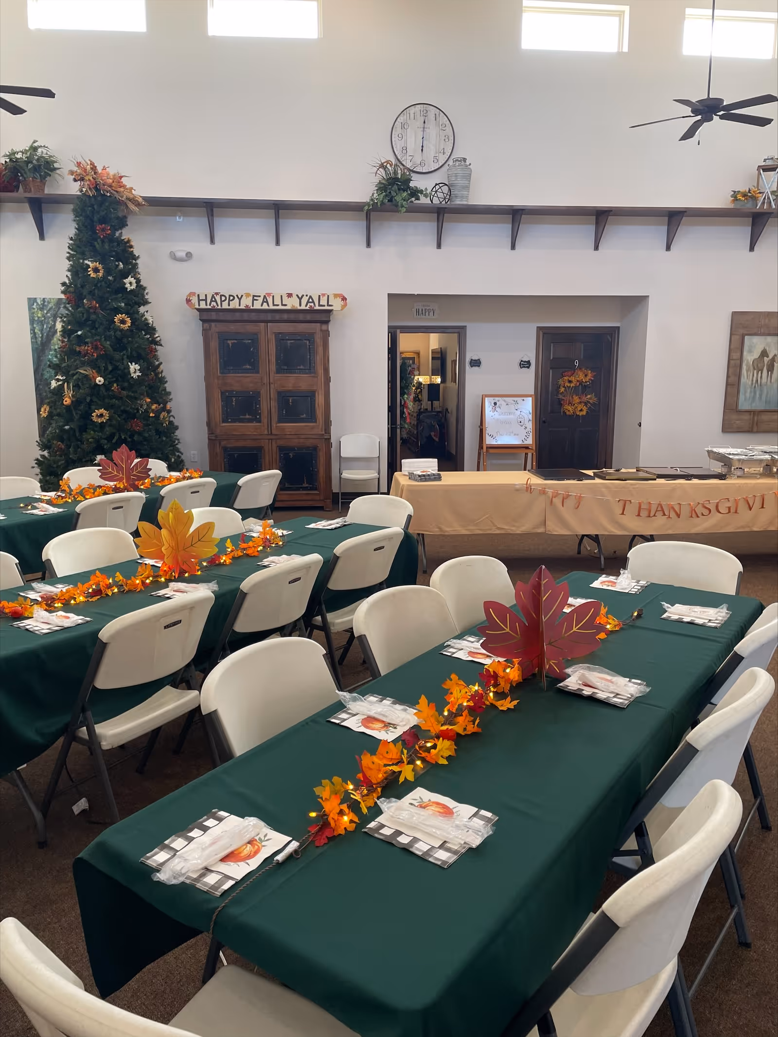 A decorated dining area with multiple tables covered in green tablecloths, set with napkins and utensils. The tables have autumn-themed centerpieces with colorful fall leaves and paper decorations. In the background, there is a tall decorated tree, a wooden cabinet, and a sign that reads 'HAPPY FALL Y'ALL'. A large clock is mounted on the wall above the cabinet, and a long table covered with a beige cloth is set up against the wall with a partially visible 'THANKSGIVING' banner. The room has high windows and ceiling fans.