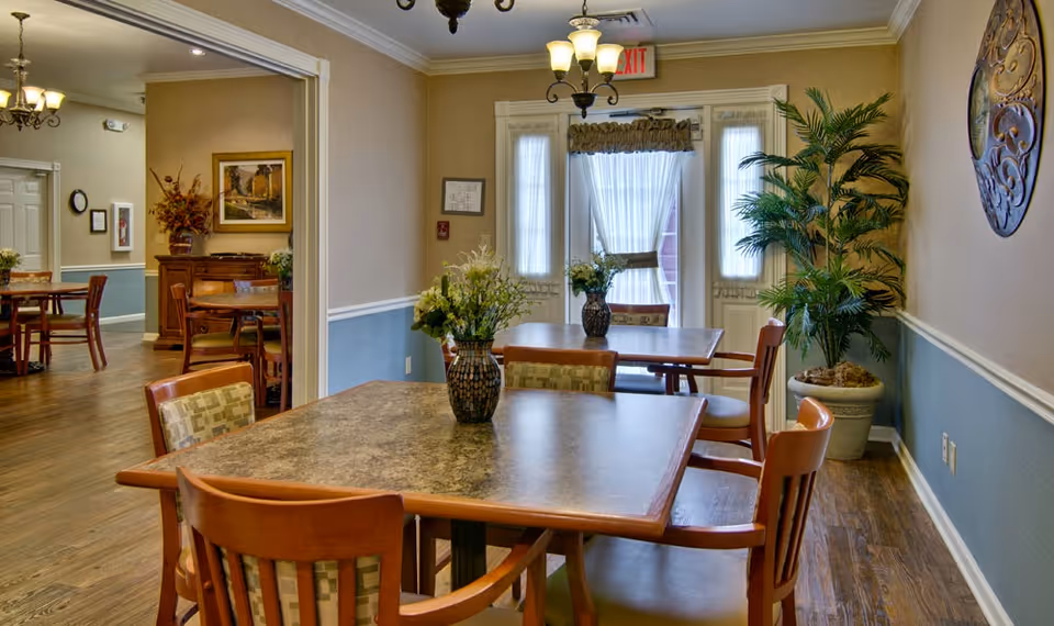 A cozy dining area in a senior living facility with wooden tables and chairs. Each table has a decorative vase with flowers. The room features wood flooring, beige and blue walls with white trim, a large potted plant in the corner, and a decorative wall piece. There is an exit door with sheer curtains letting in natural light, and another dining area is visible through an open doorway.