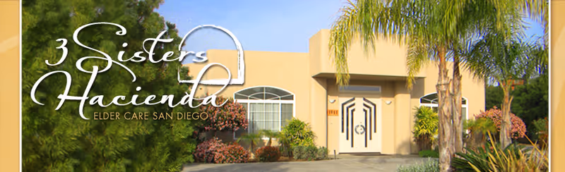 Exterior view of a single-story elder care facility building with a beige facade, decorative double front doors, and surrounding greenery including palm trees and bushes under a clear blue sky.