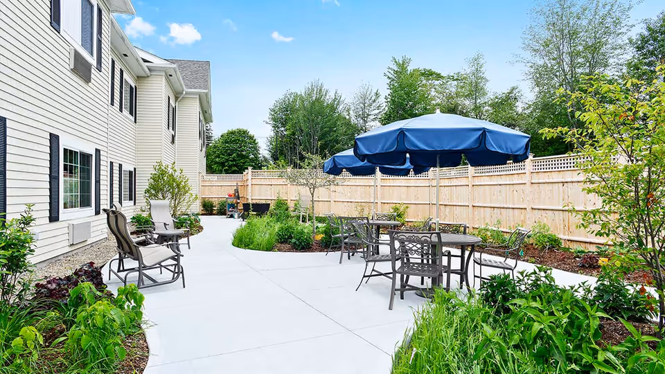Sunlit outdoor patio with tables, chairs and blue umbrellas beside a residential building and landscaped gardens.
