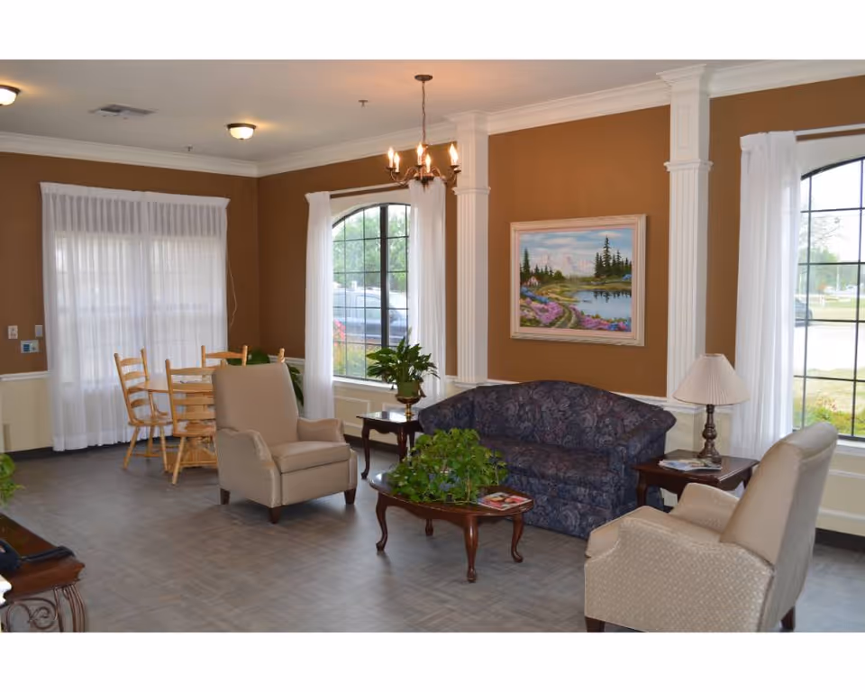 A cozy living room area with two beige armchairs and a dark patterned sofa arranged around a wooden coffee table with a green plant. There is a side table with a lamp next to the sofa. The walls are painted brown with white trim and columns, and a landscape painting hangs on the wall. Large windows with white curtains let in natural light. In the background, there is a wooden dining table with chairs near another window.