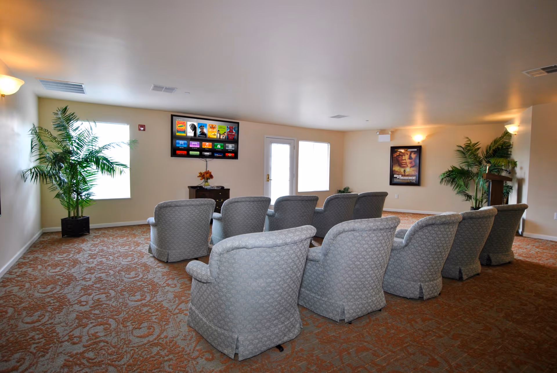 A small theater-style room with two rows of upholstered armchairs facing a wall-mounted flat screen TV. The room has beige walls, patterned carpet, two large potted plants, and a framed movie poster on the wall. There are windows and a door letting in natural light.