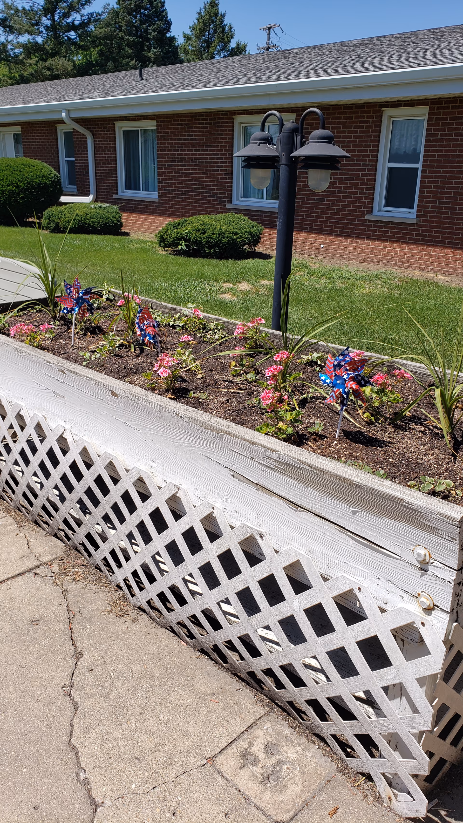A raised garden bed with pink flowers and patriotic pinwheels in front of a brick building with windows and neatly trimmed bushes. A black double-lamp post is situated behind the garden bed on a grassy lawn.