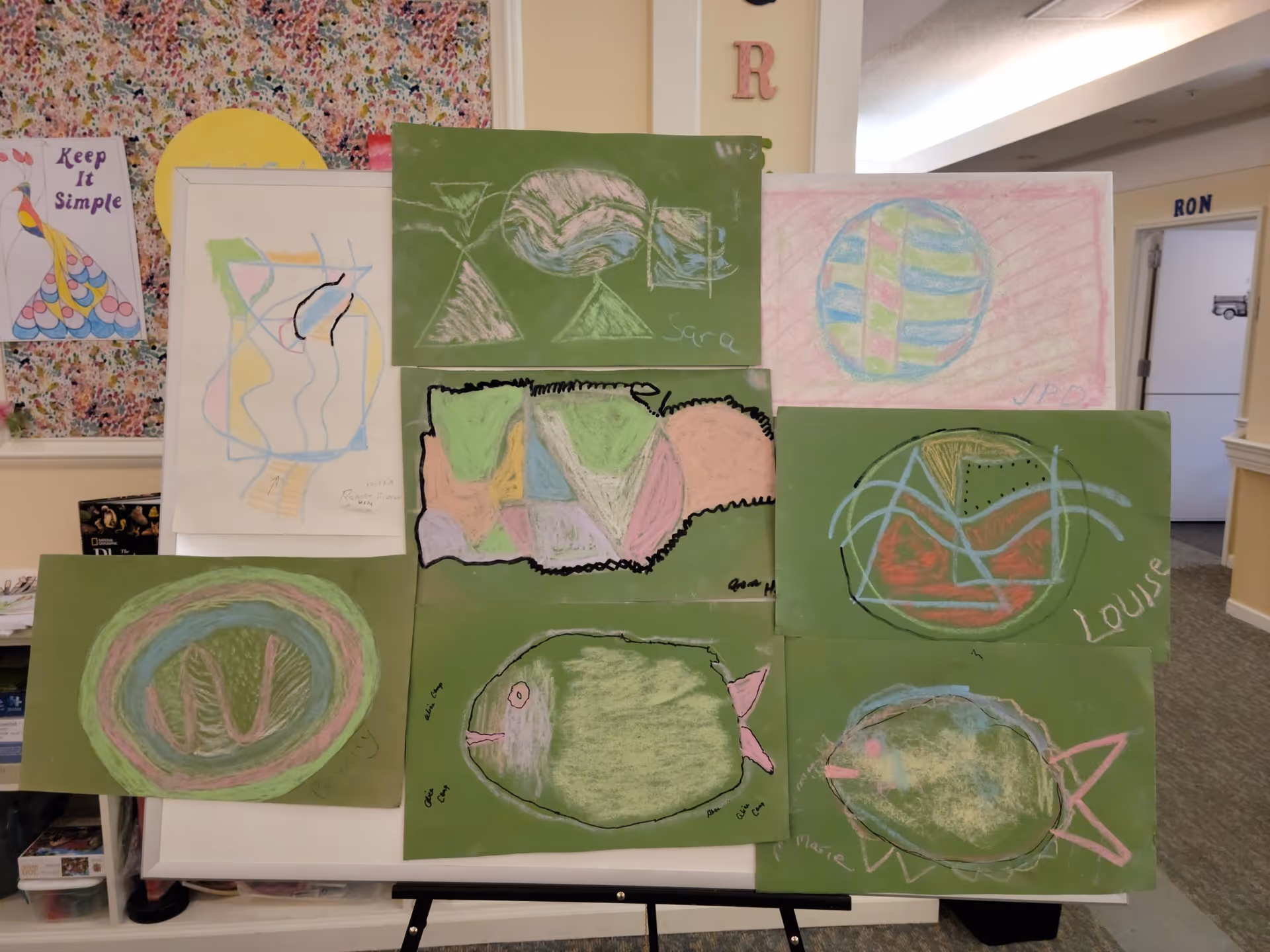 A display of colorful pastel chalk drawings on green paper arranged on an easel in a senior living facility common area.