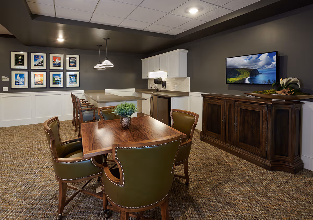 Communal dining area with a wooden table and green leather chairs, a small kitchenette, wall-mounted TV and framed artwork on the wall.