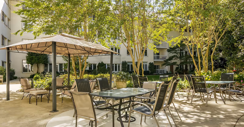 Sunlit courtyard patio with metal tables and chairs and a gazebo in front of a multi-story building.