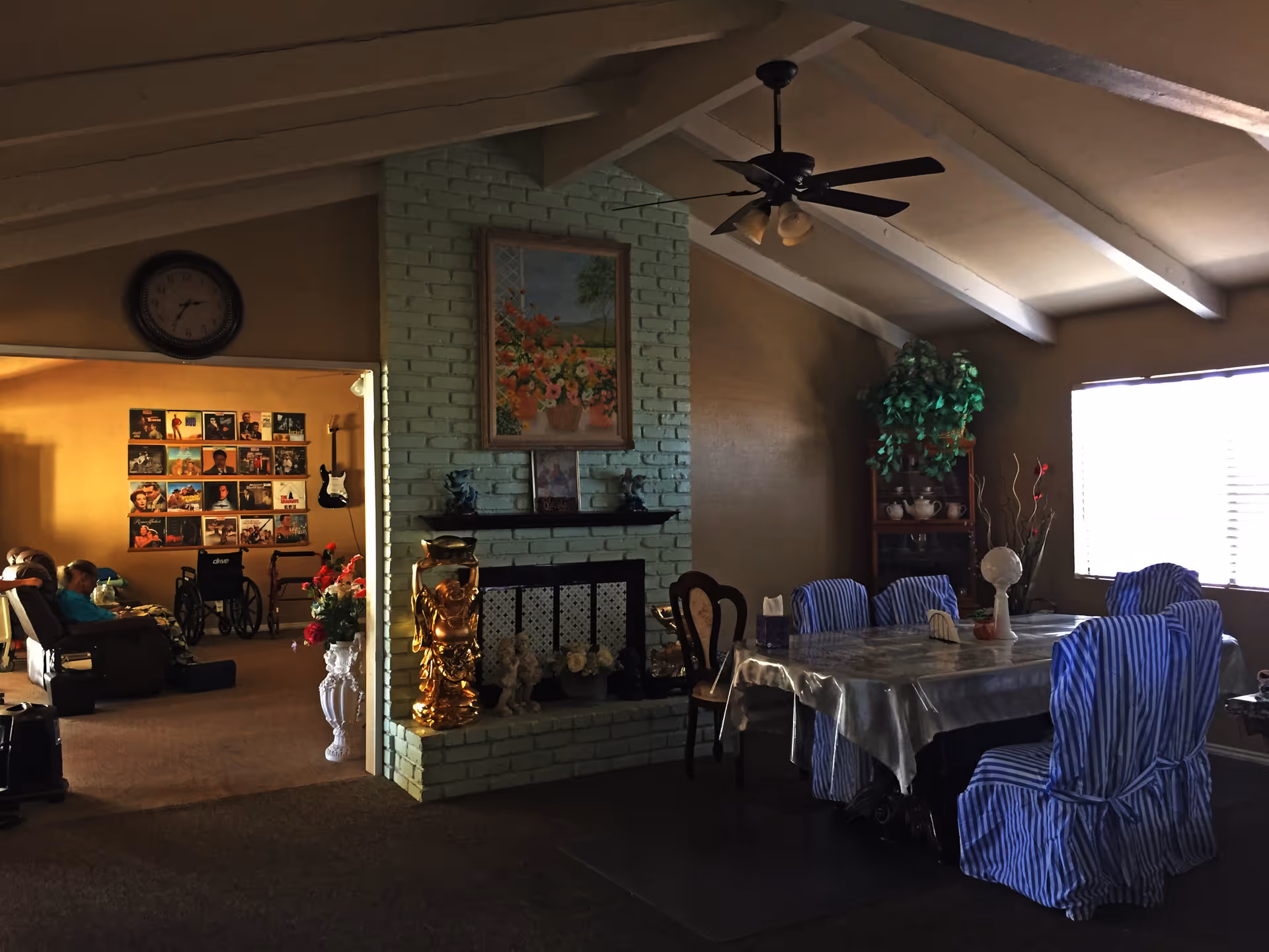 Interior view of a senior living facility showing a dining area with a table covered by a plastic tablecloth and six chairs with blue and white striped covers. A ceiling fan hangs above. To the left, there is a green painted brick fireplace with decorative items and a floral painting above it. In the adjacent room, a person is seated in a recliner near a wall decorated with framed pictures and a guitar hanging. A wheelchair and a walker are also visible in the adjacent room.