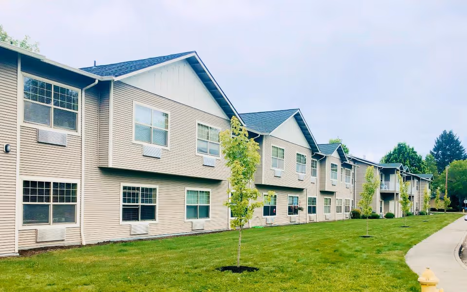 Two-story beige senior living building with multiple windows facing a grassy lawn and young trees.