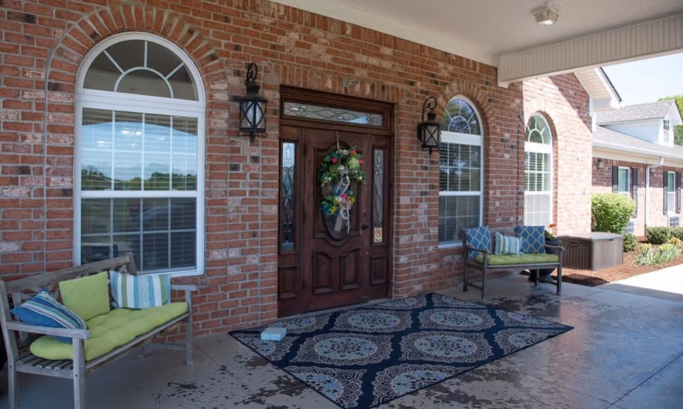 Covered entrance area of a brick building with a wooden front door decorated with a floral wreath. Two arched windows with white blinds flank the door. Two benches with colorful cushions are placed on either side of the door, and a patterned outdoor rug lies on the concrete floor. Two black wall-mounted lanterns are above the benches.