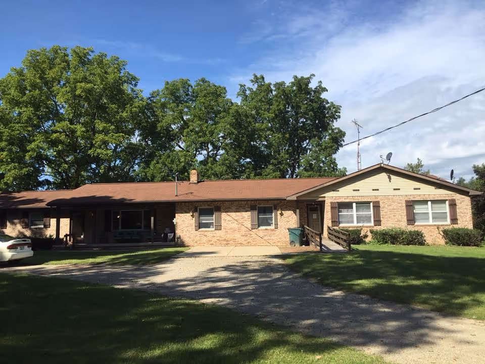 Single-story brick building with a brown roof, several windows with shutters, a small porch with a ramp, surrounded by green grass and large trees under a partly cloudy blue sky.