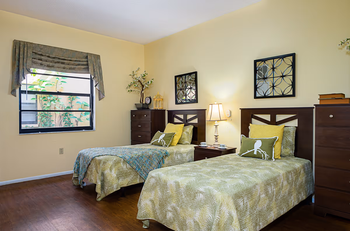 A bright bedroom with two twin beds featuring green patterned bedspreads and decorative pillows. Between the beds is a wooden nightstand with a lamp and two cups. The room has light yellow walls, two framed decorative mirrors above the beds, a window with a valance, and two dark wooden dressers with decorative items on top.